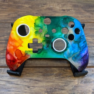 May include: A custom-painted video game controller with a rainbow-colored design. The controller is on a black stand and has white buttons.