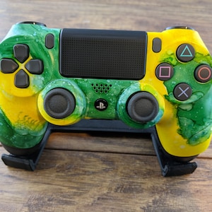 May include: A custom-painted video game controller with a green and yellow marbled design. The controller has black buttons and joysticks, and is resting on a black stand. The PlayStation logo is visible on the controller.