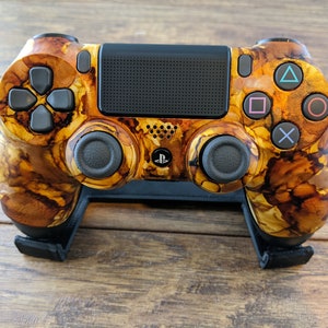 May include: A custom-painted PlayStation 4 controller with a brown and gold marbled design. The controller is on a black stand.