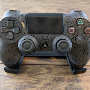 May include: A black PlayStation 4 controller with a speckled, textured finish. The controller is on a black stand and features the standard PlayStation buttons: triangle, square, circle, and X.