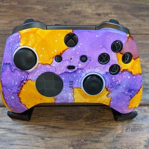 May include: An Xbox controller with a custom design. The controller features a vibrant abstract pattern in shades of purple and yellow, with white speckles. The buttons and joysticks are black.