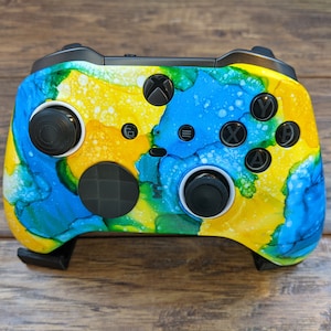 May include: A custom Xbox controller with a vibrant abstract design in yellow, blue, and green. The controller features black buttons, thumbsticks, and triggers. The design resembles an ink-blot effect, adding a unique aesthetic to the gaming accessory.