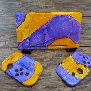 May include: A purple and yellow Nintendo Switch console and two matching controllers. The console and controllers are decorated with a marbled paint design.