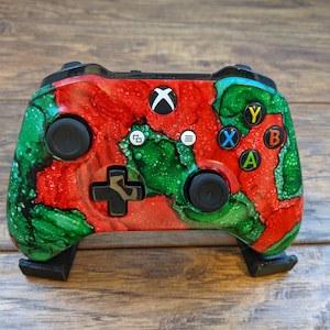 May include: A black Xbox controller with a red and green abstract design. The controller has a black Xbox logo on the top and black buttons.
