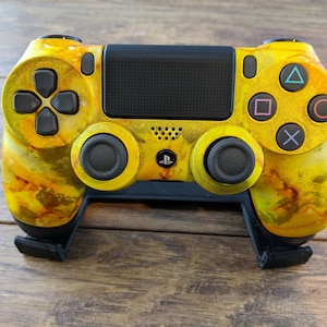 May include: A yellow and orange PlayStation 4 controller with black buttons and a black touchpad. The controller is sitting on a black stand.