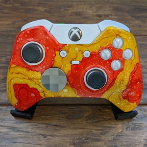 May include: A white Xbox controller with a custom red, yellow, and white paint job. The controller has a black faceplate and black buttons.