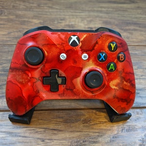 May include: A red and black Xbox controller with a marbled design. The controller has a black D-pad, black buttons, and a black Xbox button.