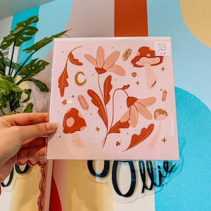 May include: A pink print with a whimsical illustration of flowers, mushrooms, and crystals. The print has a gold crescent moon and stars. The text "Jollie Tillie Print" is in the top right corner.