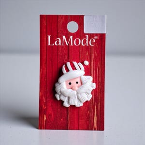 May include: A Santa Claus-shaped embellishment with a white beard, hat, and pink face, attached to a red wooden background. The embellishment has a glittery trim. The brand name "LaMode" is printed on the red background.