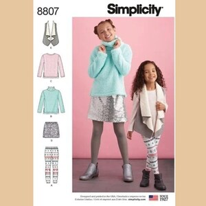 May include: A Simplicity sewing pattern featuring children's clothing. The image shows two girls wearing outfits made from the pattern. One girl wears a turquoise turtleneck sweater and a silver sequin skirt. The other wears a white sweater, gray vest, and patterned leggings.