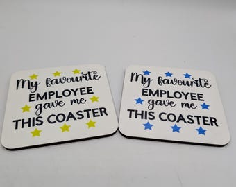 May include: Two white square coasters with black text that reads "My favourite EMPLOYEE gave me THIS COASTER." Yellow and blue stars accent the text. The coasters are on a white surface.