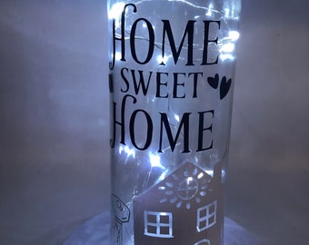 May include: A clear glass bottle with a string of white lights inside. The bottle has a black vinyl decal that says "Home Sweet Home" with two hearts. The bottle also has a black vinyl decal of a house with a key.