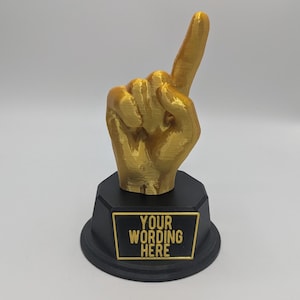 May include: A gold-colored 3D printed hand sculpture with the index finger pointing upward, mounted on a black octagonal base. A rectangular plaque on the front of the base features the raised gold text "YOUR WORDING HERE" against a black background.