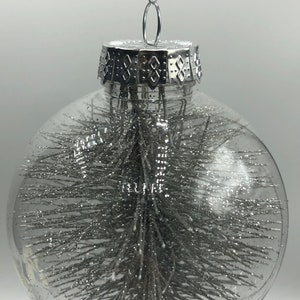 May include: A clear spherical glass ornament featuring a silver glitter-covered pine tree inside. The ornament is topped with a silver metallic cap and includes a thin silver string for hanging, creating a reflective and textured appearance.
