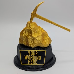 May include: A gold-colored pickaxe rests atop a gold-colored rock formation, mounted on a black base with a gold-bordered plaque. The plaque reads "YOUR WORDING HERE". The trophy is set against a white background.