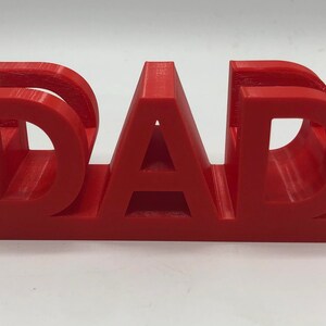 May include: A bright red, three-dimensional plastic object featuring the word DAD in bold, capital letters. The letters are connected at the base, creating a sturdy, freestanding structure with visible horizontal layer lines from the 3D printing process.