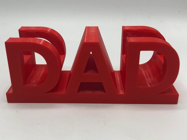 May include: A bright red, three-dimensional plastic object featuring the word DAD in bold, capital letters. The letters are connected at the base, creating a sturdy, freestanding structure with visible horizontal layer lines from the 3D printing process.