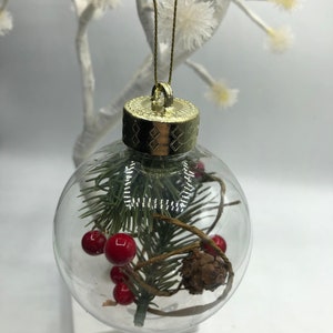 May include: A clear, spherical glass ornament featuring a gold-colored cap and a gold hanging string. Inside the hollow sphere are green artificial pine needles, small red berries, a brown pinecone, and thin brown twine wrapped around the foliage.
