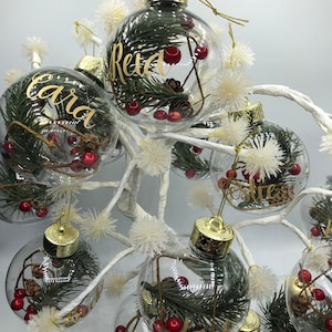 May include: Clear spherical Christmas ornaments with gold caps, each containing green pine needles, small brown pinecones, and red berries. The exterior of each glass bauble features a custom name written in elegant gold cursive lettering.