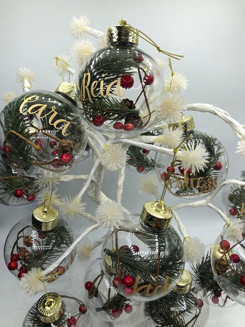 May include: Clear spherical Christmas ornaments with gold caps, each containing green pine needles, small brown pinecones, and red berries. The exterior of each glass bauble features a custom name written in elegant gold cursive lettering.