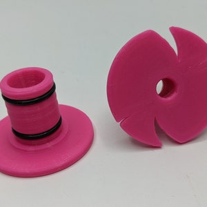 May include: Two pink plastic 3D printed objects. One is a round disc with a hole in the centre and five points. The other is a cylinder with a round base and a ring around the top.