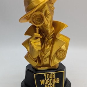 May include: A gold-coloured bust of a detective wearing a fedora and trench coat, holding a magnifying glass to one eye. The figure stands on a black octagonal base featuring a rectangular plaque with the text "YOUR WORDING HERE" in gold letters.
