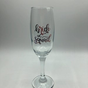 May include: A clear glass champagne flute with the words "bride squad" written in gold lettering.