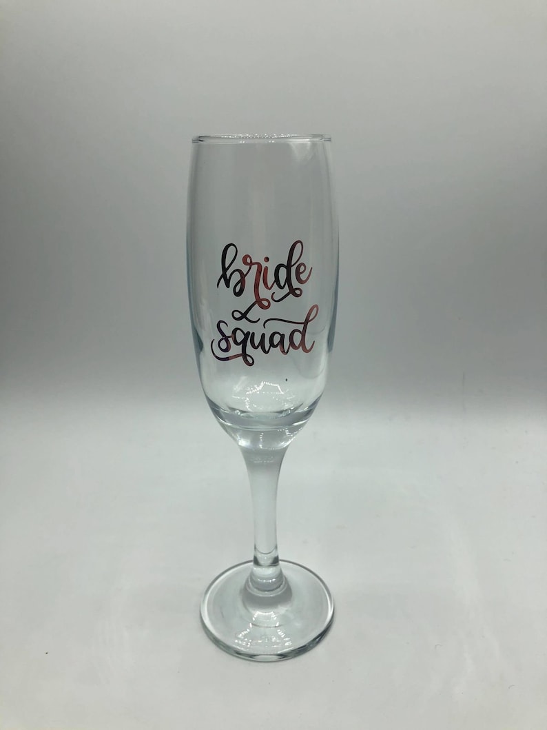 May include: A clear glass champagne flute with the words "bride squad" written in gold lettering.