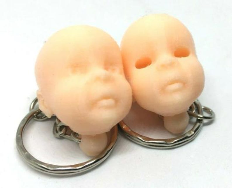 May include: Two small, realistic-looking baby doll heads attached to keychains. The dolls have a light peach skin tone and are made of a smooth, soft material.