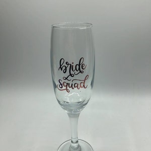 May include: A clear glass champagne flute with the text "bride squad" in black script on the side.