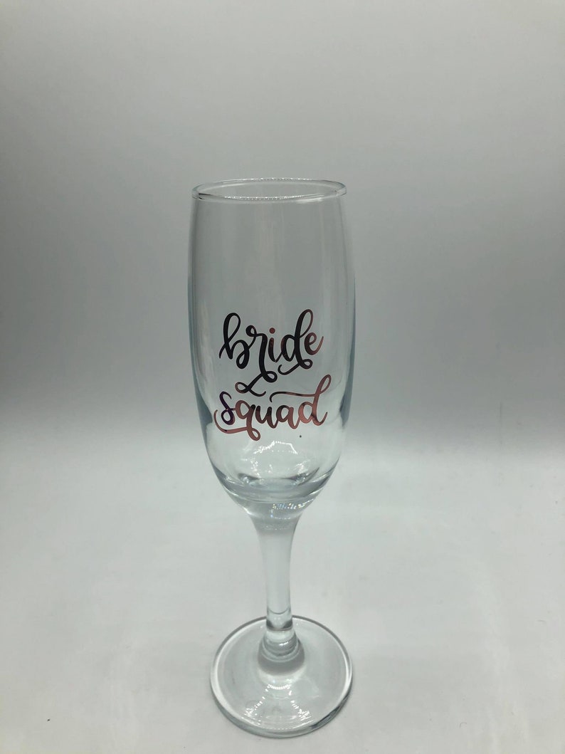 May include: A clear glass champagne flute with the text "bride squad" in black script on the side.