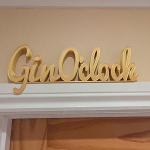 May include: Gold-colored wooden sign with the text "Gin O'clock" in a cursive font.
