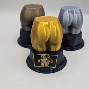 May include: Three trophies featuring a sculpted human posterior in metallic gold, silver, and bronze finishes. Each figure is mounted on a black hexagonal base with a plaque that reads YOUR WORDING HERE in gold lettering on the front-most trophy.