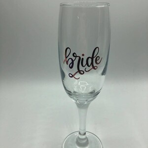 May include: A clear glass champagne flute with the word "bride" written in gold lettering.
