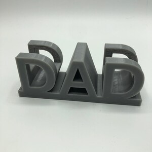May include: A three-dimensional gray plastic object featuring the word DAD in block capital letters. The letters are connected at the base, creating a sturdy, freestanding structure with visible horizontal layer lines from the manufacturing process.