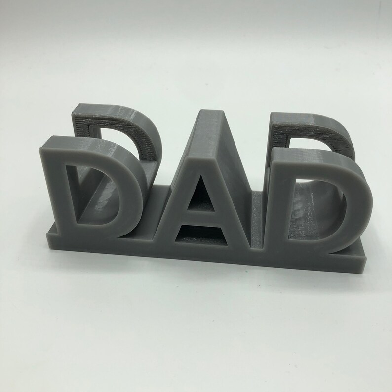 May include: A three-dimensional gray plastic object featuring the word DAD in block capital letters. The letters are connected at the base, creating a sturdy, freestanding structure with visible horizontal layer lines from the manufacturing process.