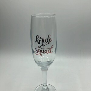 May include: A clear glass champagne flute with the text "bride squad" in black and rose gold lettering.