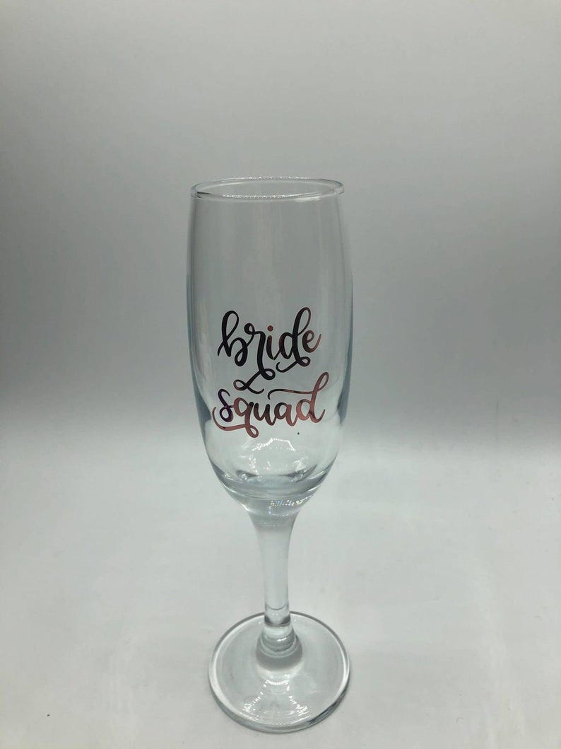 May include: A clear glass champagne flute with the text "bride squad" in black and rose gold lettering.