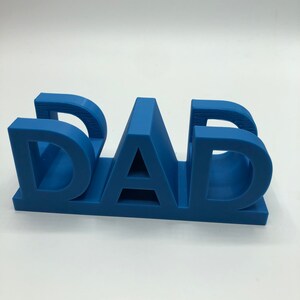 May include: A three-dimensional blue plastic sign featuring the word DAD in bold, block capital letters. The letters are connected at the base, creating a sturdy, freestanding structure with a smooth matte finish and visible horizontal print lines.