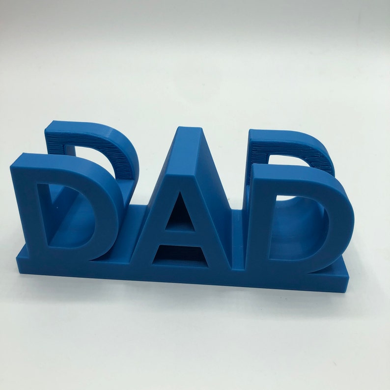 May include: A three-dimensional blue plastic sign featuring the word DAD in bold, block capital letters. The letters are connected at the base, creating a sturdy, freestanding structure with a smooth matte finish and visible horizontal print lines.