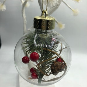 May include: A clear spherical Christmas ornament featuring a gold-colored cap and a gold hanging string. Inside the transparent globe are green pine needles, small red berries, a brown pinecone, and thin brown decorative twine.