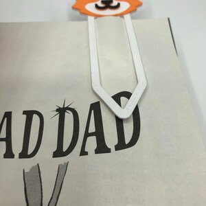 May include: White bookmark with a 3D printed orange and white corgi head. The bookmark is shaped like a traditional bookmark with a pointed end. The corgi has black eyes and a black nose.