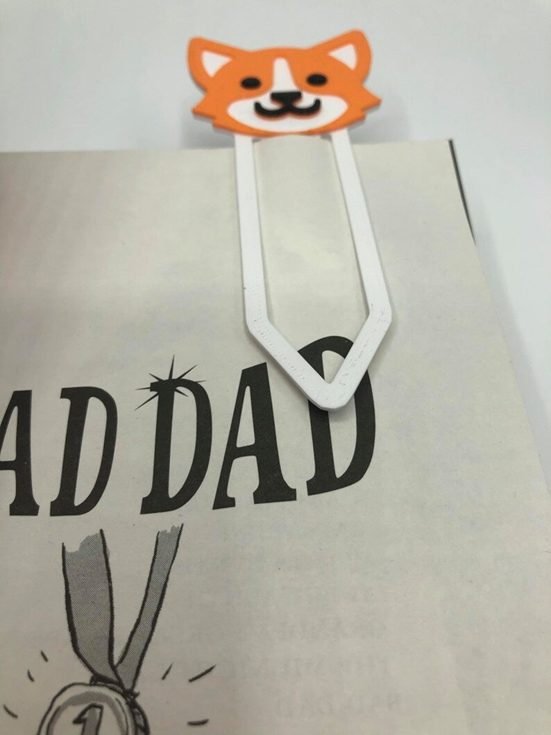 May include: White bookmark with a 3D printed orange and white corgi head. The bookmark is shaped like a traditional bookmark with a pointed end. The corgi has black eyes and a black nose.
