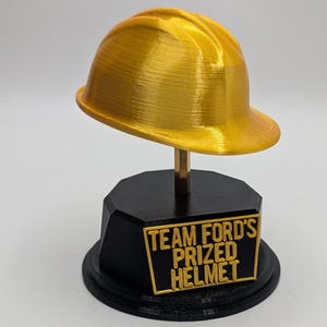 May include: A gold-coloured hard hat trophy mounted on a black octagonal base. The base features a gold-coloured rectangular plaque with the raised text "TEAM FORD'S PRIZED HELMET" in capital letters. The hard hat has a smooth, glossy finish.