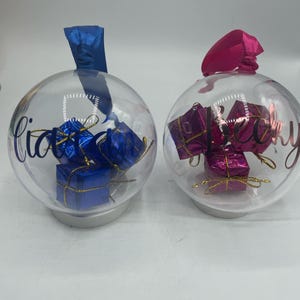 May include: Two clear spherical ornaments with silver bases and hanging ribbons. The left ornament has a blue ribbon and contains blue gift boxes, with the name Liam written in blue. The right ornament has a pink ribbon, pink gift boxes, and the name Becky.