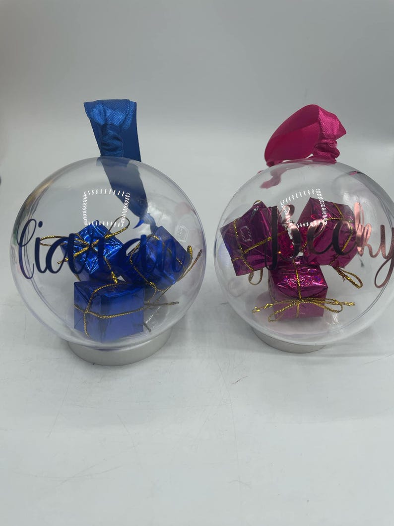 May include: Two clear spherical ornaments with silver bases and hanging ribbons. The left ornament has a blue ribbon and contains blue gift boxes, with the name Liam written in blue. The right ornament has a pink ribbon, pink gift boxes, and the name Becky.