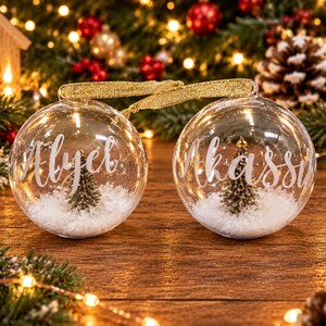 May include: Two clear spherical glass ornaments featuring white script text reading Alyet and Akassia. Each ornament contains a miniature green pine tree set in artificial white snow and is connected by a gold ribbon for hanging.