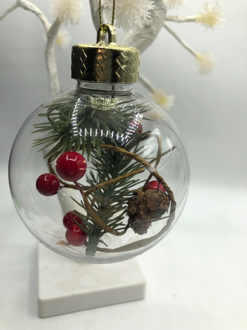 May include: A clear, spherical Christmas ornament featuring a gold-colored cap and a gold hanging string. Inside the transparent globe are green pine needles, small red berries, a brown pinecone, and a thin, twisted brown vine-like element.