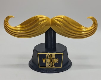 May include: A gold-coloured moustache sculpture mounted on a black pedestal base. The base features a rectangular plaque with the text "YOUR WORDING HERE" in gold lettering. The moustache has a textured, curved design with pointed ends.