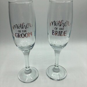 May include: Two clear glass champagne flutes with gold lettering. The left flute says "Mother of the Groom" and the right flute says "Mother of the Bride".
